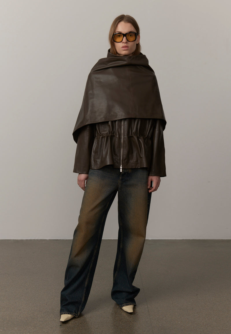 A model wearing a dark brown leather cape-style jacket with a high draped collar, paired with dark gradient wide-leg jeans and yellow-tinted sunglasses, from Herskind. 