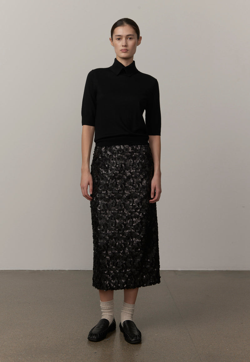 A model stands wearing a black short-sleeve knit polo-style top and a black patterned midi skirt, paired with white socks and black loafers from Herskind. 