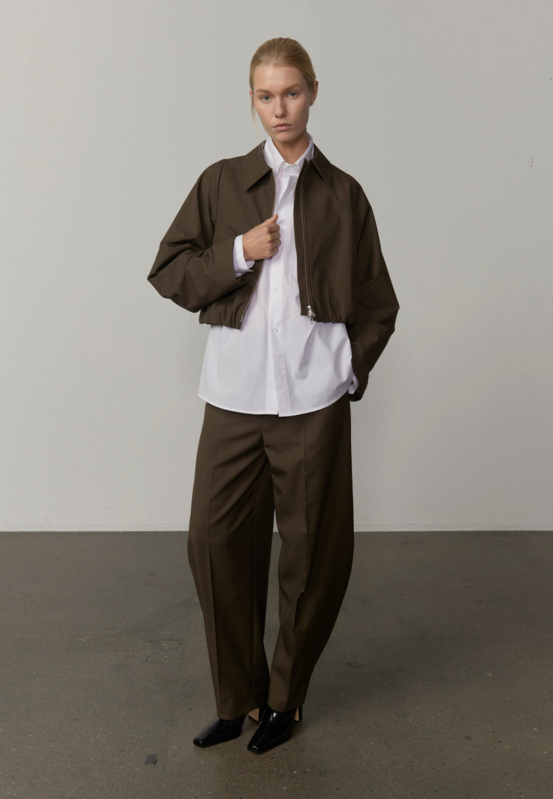 A model wearing a dark brown cropped jacket, matching dark brown trousers, a white button-up shirt, and black shoes from Herksind. 