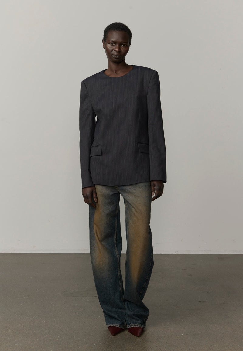 A model stands in a studio wearing a dark charcoal gray long-sleeve top with flap pockets and wide-leg faded jeans in blue and brown tones, paired with dark red pointed shoes from Herskind. 