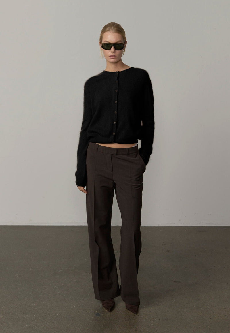 A model stands in a minimalist studio setting wearing a black cropped button-front cardigan, dark brown wide-leg trousers, and black sunglasses from Herskind. 