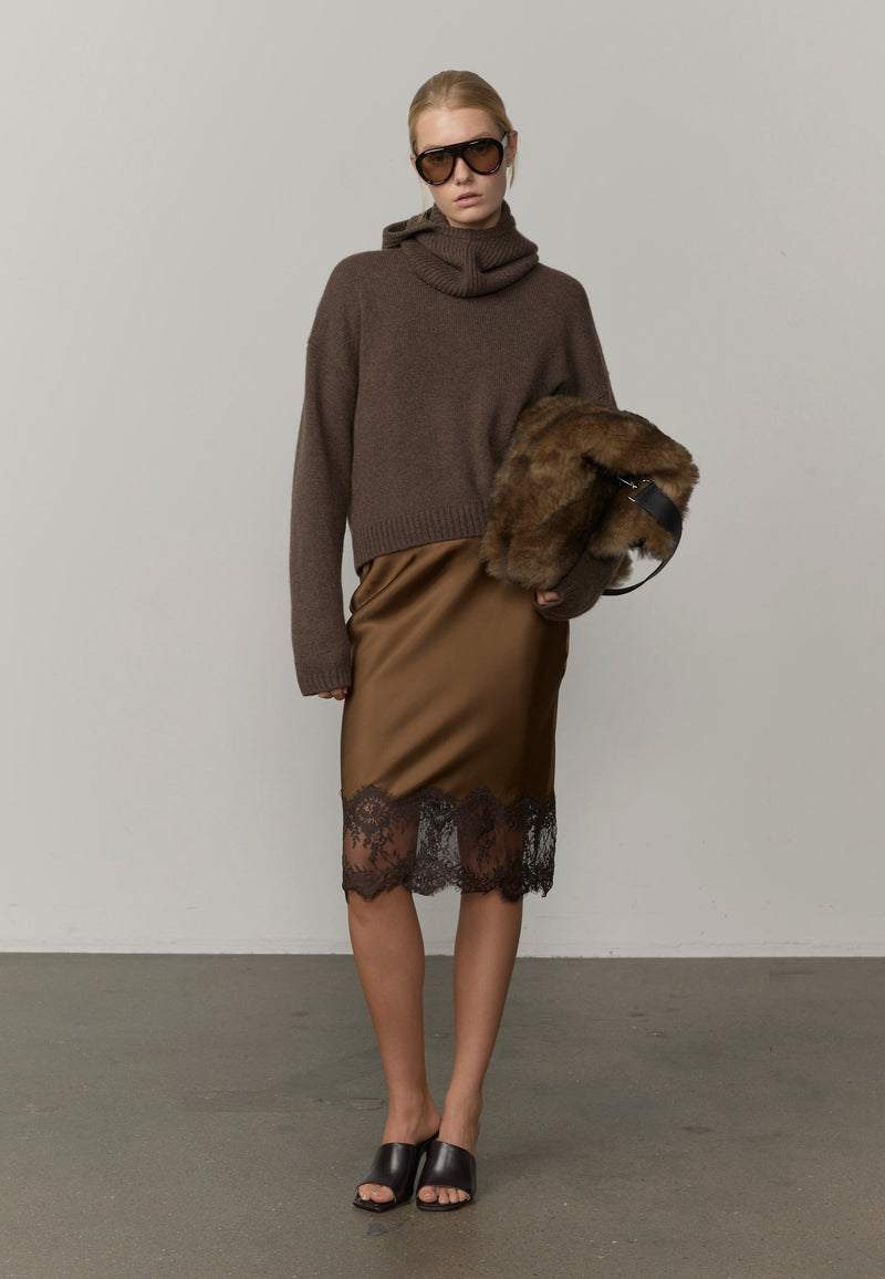 A model wearing a dark brown cowl-neck knit sweater, a brown satin pencil skirt with black lace hem, black slide sandals, and holding a brown fur-trimmed handbag from Herskind. 
