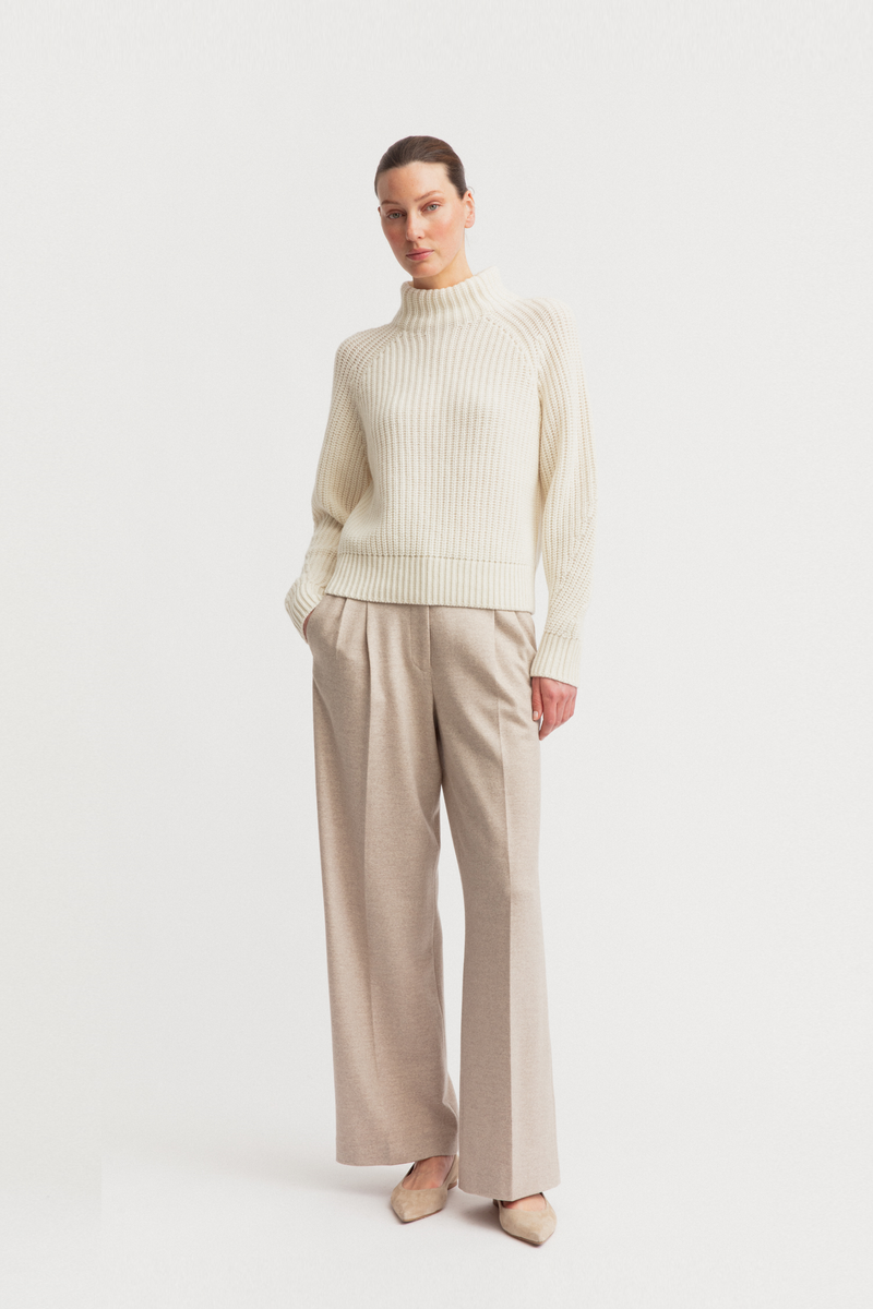 A woman modeling a cream ribbed knit mock-neck sweater and beige wide-leg trousers against a plain light gray studio background.