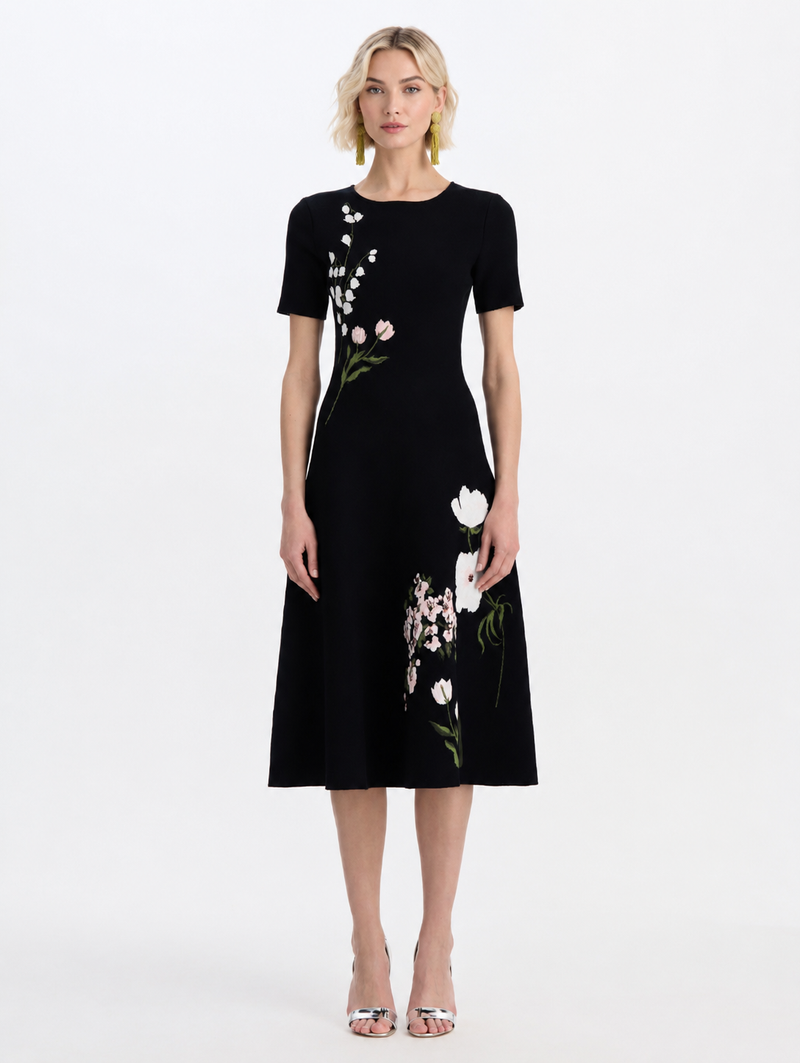 A woman modeling a black short-sleeve midi dress with white floral embroidery and green stems, by Oscar de la Renta. 