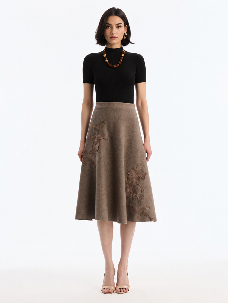 A model wearing a black short-sleeve mock-neck top and a brown A-line midi skirt with tonal floral embroidery by Oscar de la Renta. 