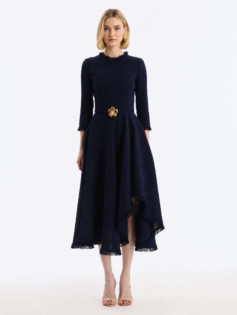 A woman modeling a navy blue midi dress with three-quarter sleeves, a ruffled high neck, and a gold floral belt at the waist, from Oscar de la Renta. 