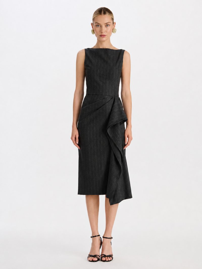 A model wearing a sleeveless black midi dress with a subtle vertical pinstripe, structured shoulders, and a draped asymmetrical skirt detail, by Oscar de la Renta. 