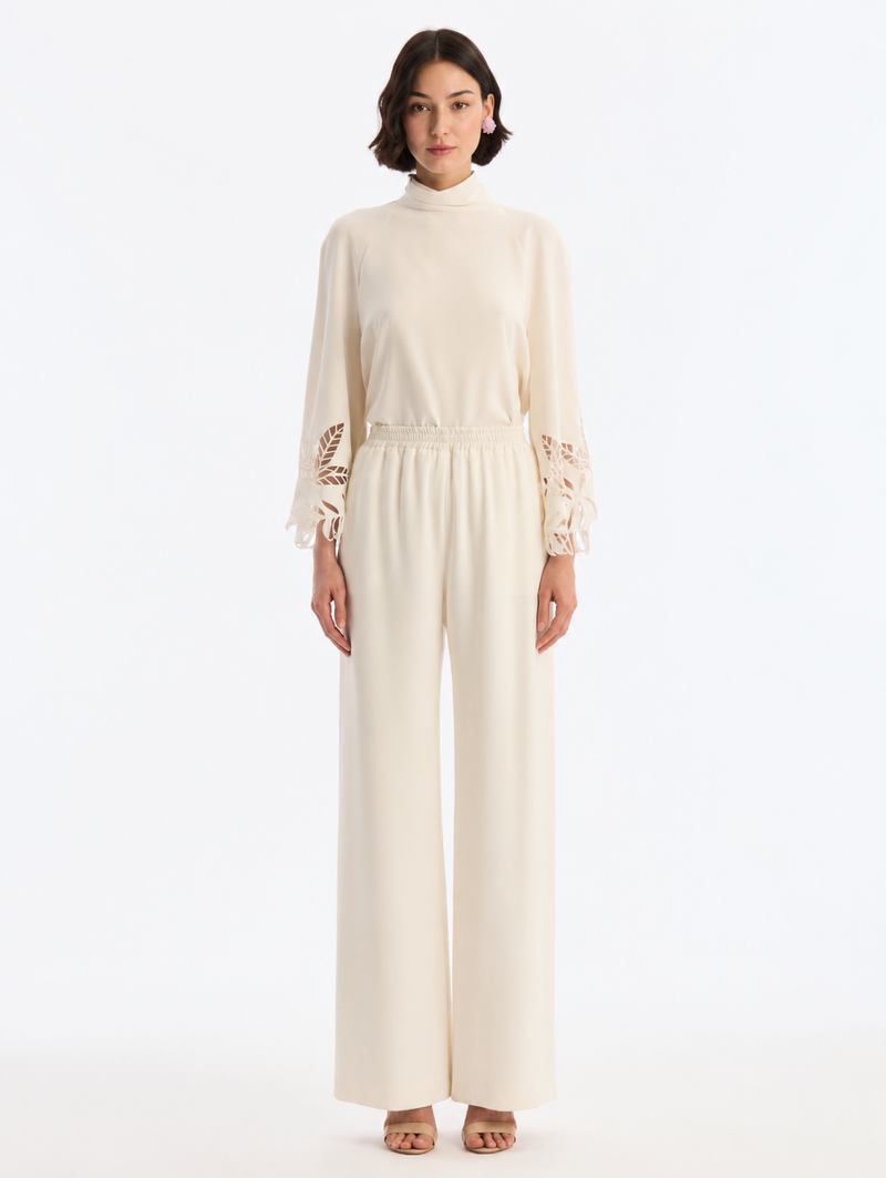 A woman modeling a cream-colored long-sleeve jumpsuit with lace detailing on the cuffs, from Oscar de la Renta. 