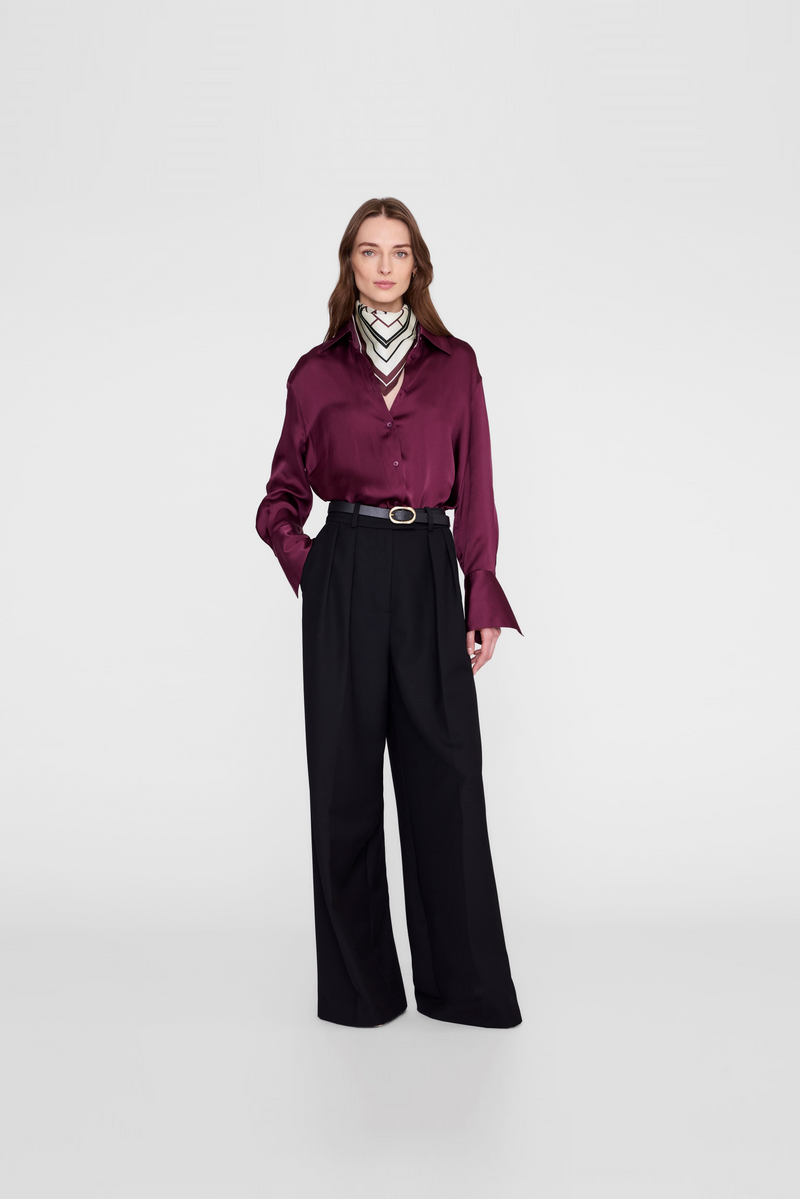 A model  wearing a long-sleeve burgundy satin shirt, black wide-leg trousers, a black belt, and a striped neck scarf by Anine Bing. 