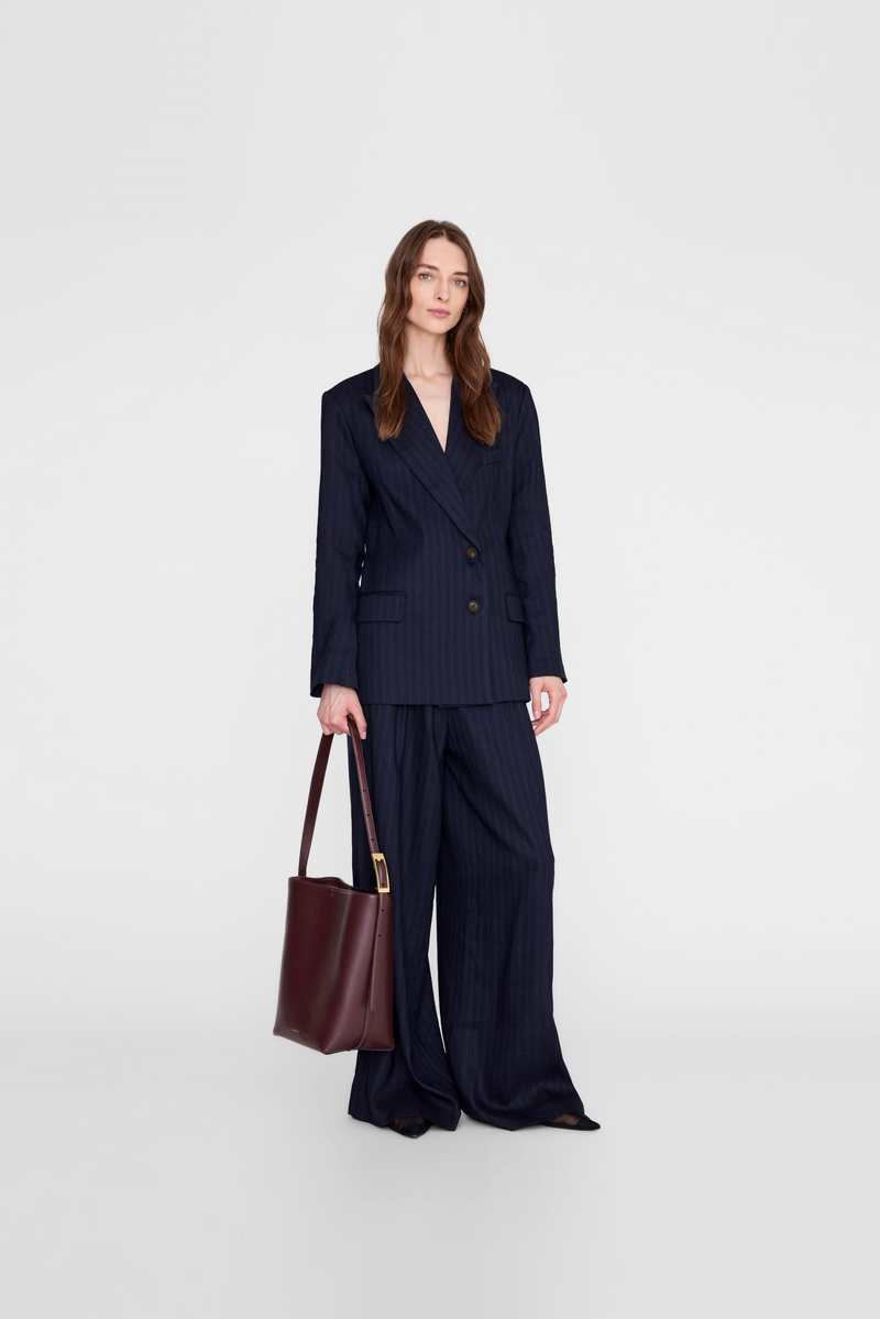 A model wearing a dark navy double-breasted blazer and matching wide-leg trousers, holding a dark burgundy leather tote bag from Anine Bing. 