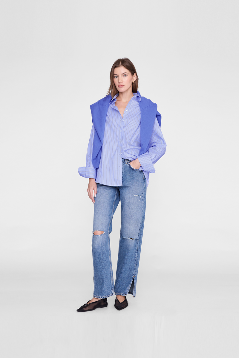 A model  wearing a light blue striped button-up shirt, a bright blue sleeveless vest, distressed blue jeans, and black pointed flats by Anine Bing. 