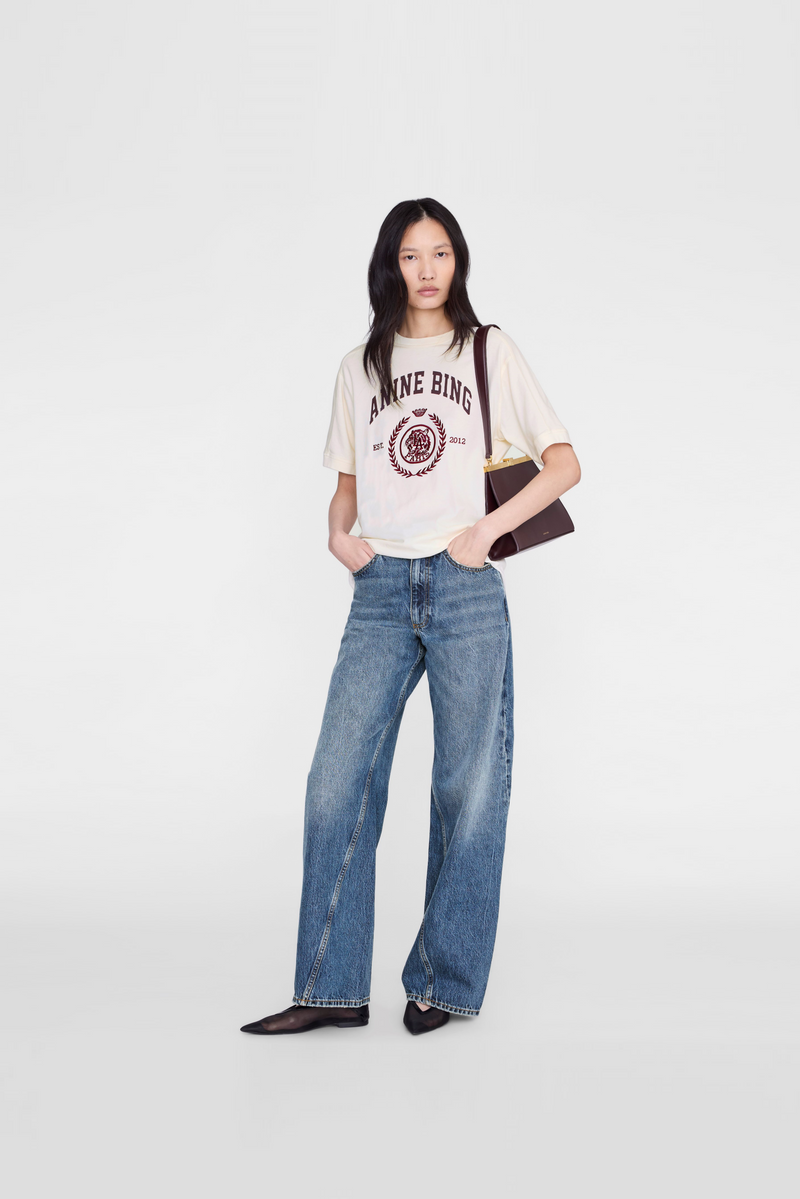 A model wearing a cream graphic t-shirt with' text, wide-leg blue jeans, and black pointed shoes by Anine Bing. 