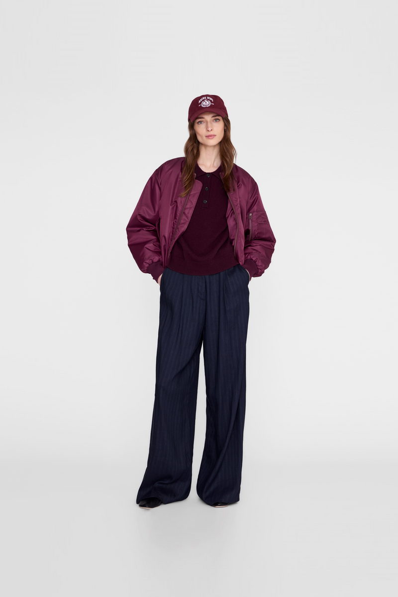 A model wearing a dark burgundy satin bomber jacket, a matching burgundy top, dark navy wide-leg trousers, and a burgundy baseball cap by Anine Bing. 