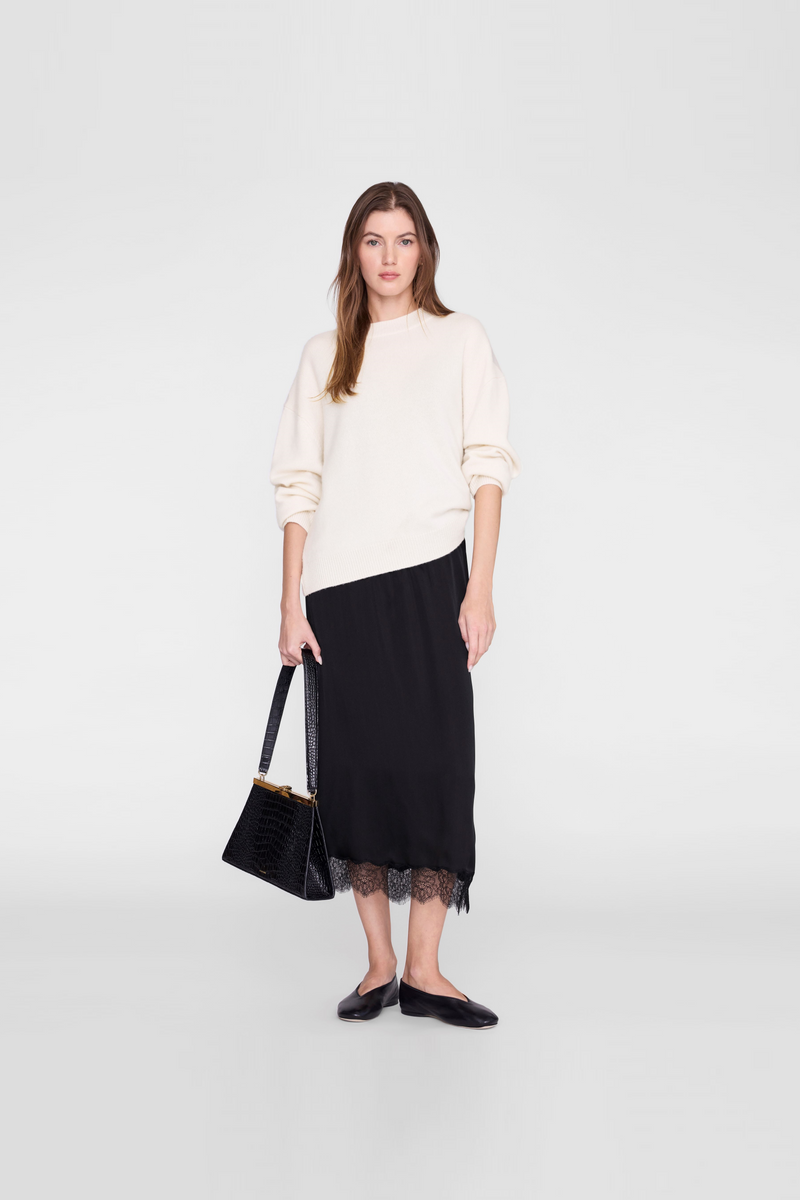 A model wearing a cream-colored asymmetrical long-sleeve top, a black midi skirt with a sheer black lace hem, black flats, and holding a black handbag by Anine Bing. 