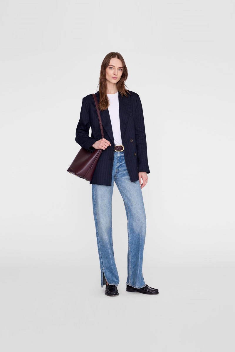 A woman wearing a dark navy blazer, light blue jeans, a white top, and black shoes, while carrying a dark burgundy shoulder bag by Anine Bing. 