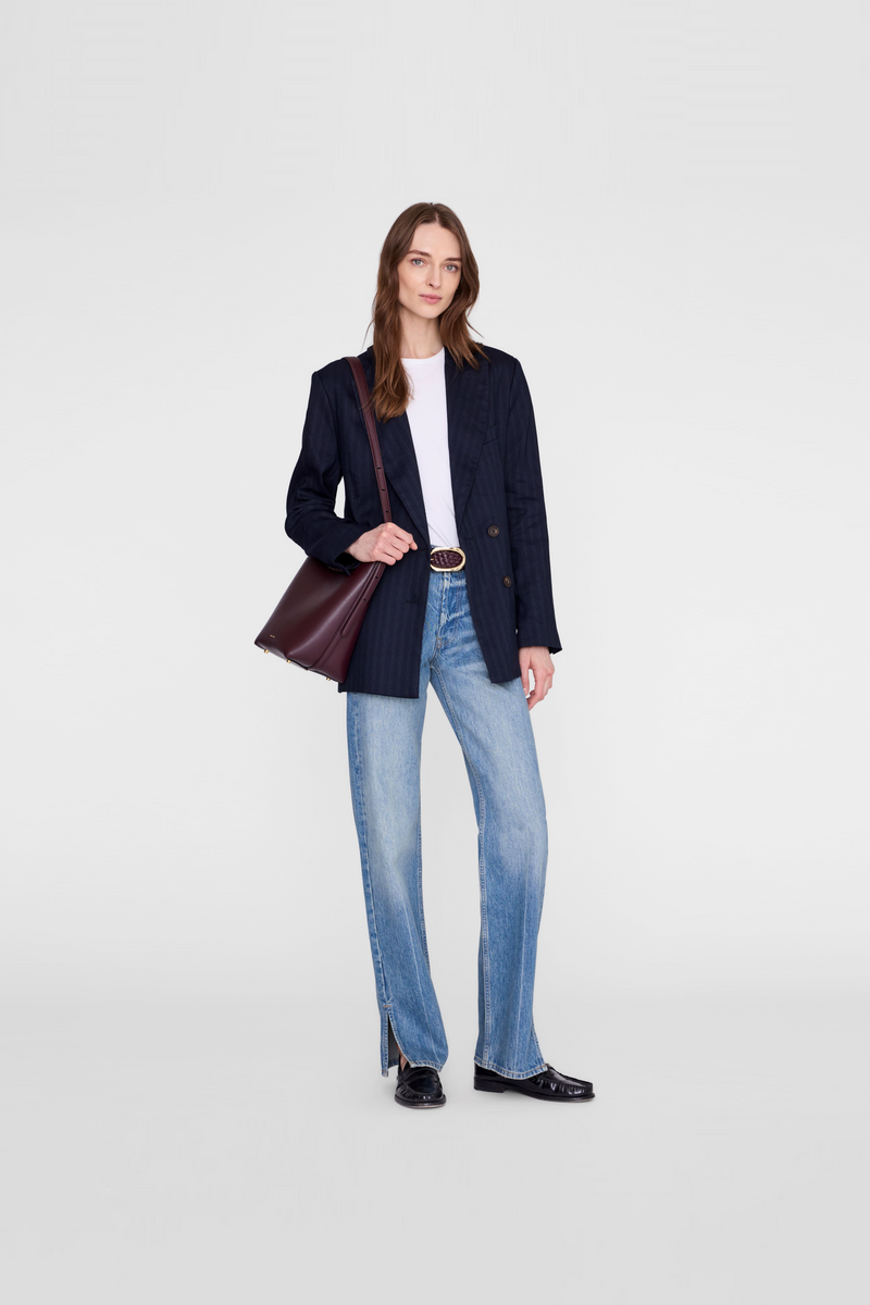 A woman wearing a dark navy blazer, light blue jeans, a white top, and black shoes, while carrying a dark burgundy shoulder bag by Anine Bing. 