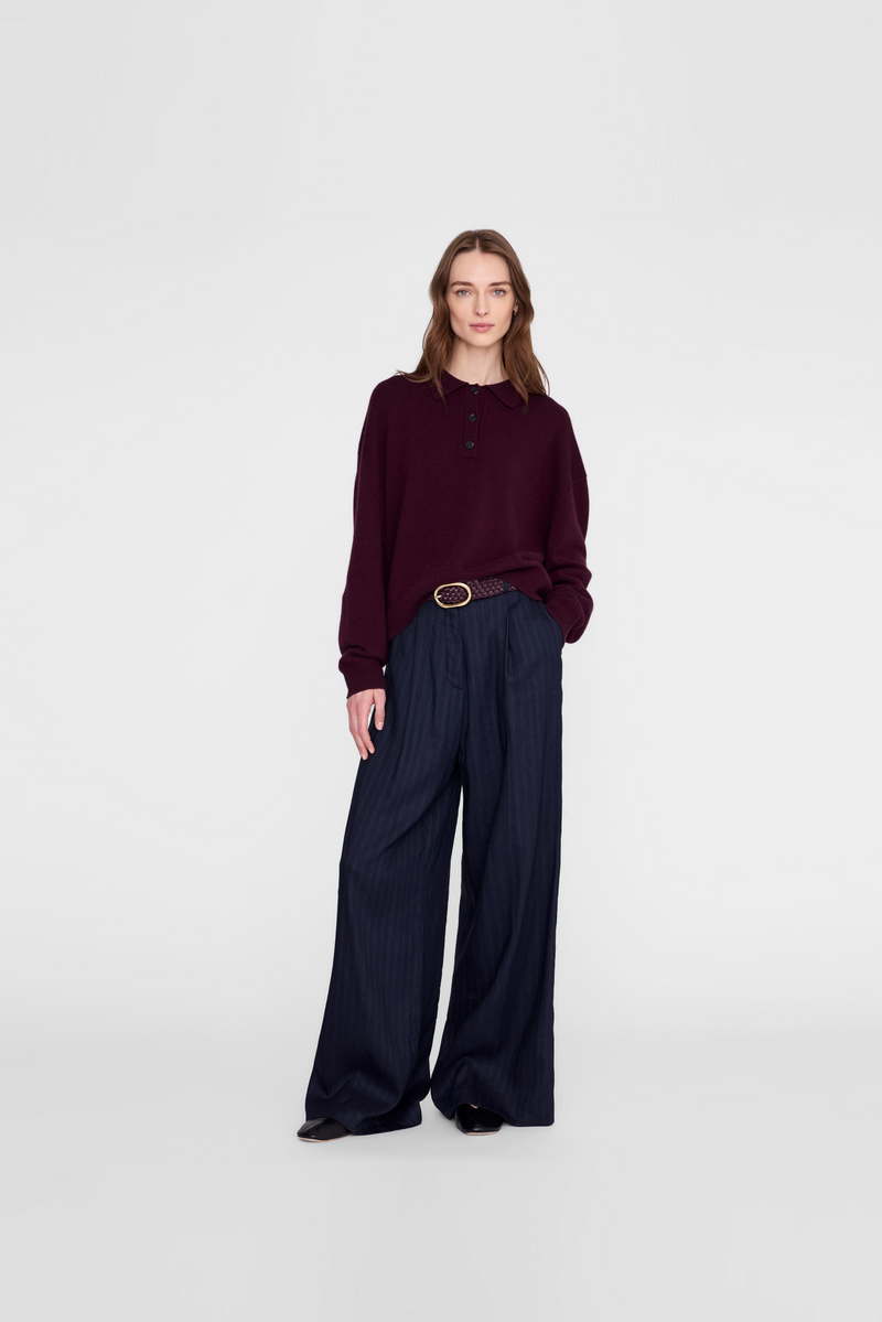 A model wearing a dark burgundy long-sleeve top and dark navy wide-leg pleated trousers with a thin gold belt by Anine Bing. 