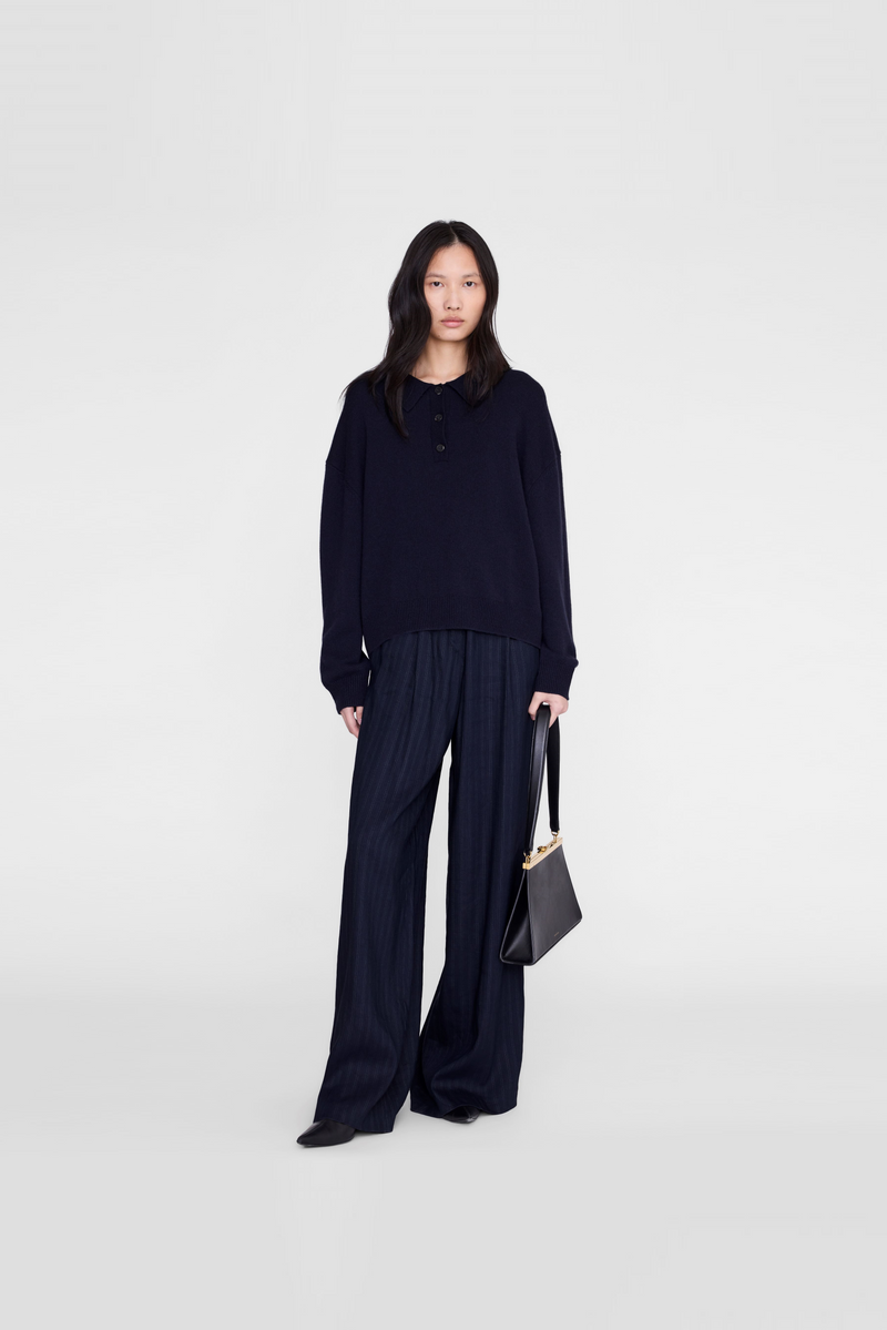 A model wearing a dark navy long-sleeve knit top and matching wide-leg pleated trousers, holding a black handbag by Anine Bing. 
