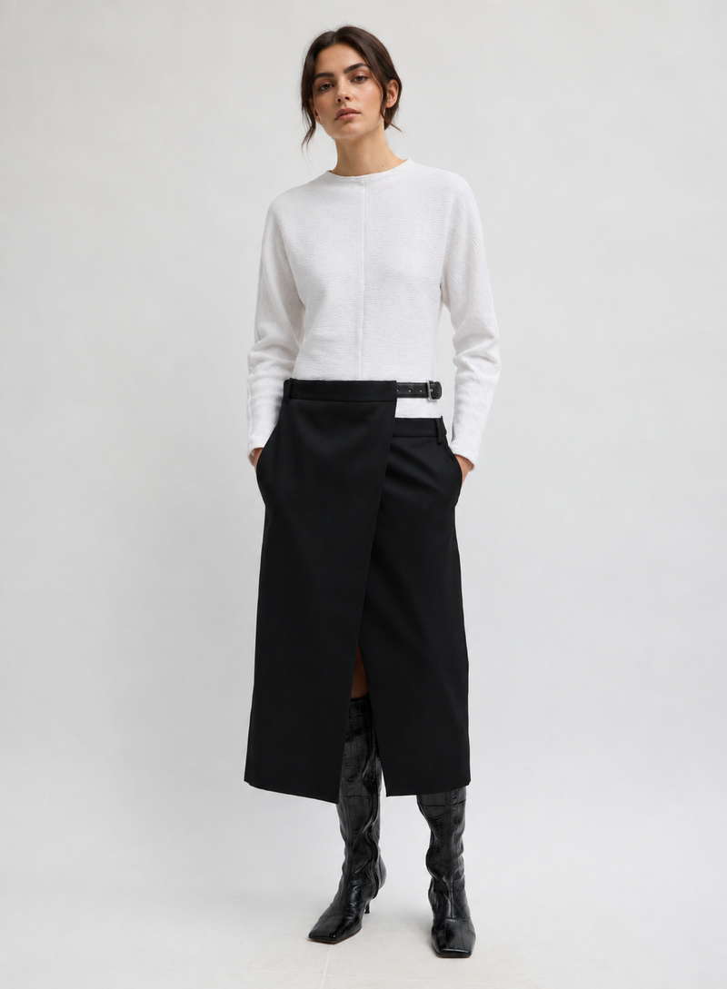 A model wearing a white long-sleeve knit top, a black wrap-style midi skirt with a front slit and black belt, and black heeled ankle boots by Tibi. 