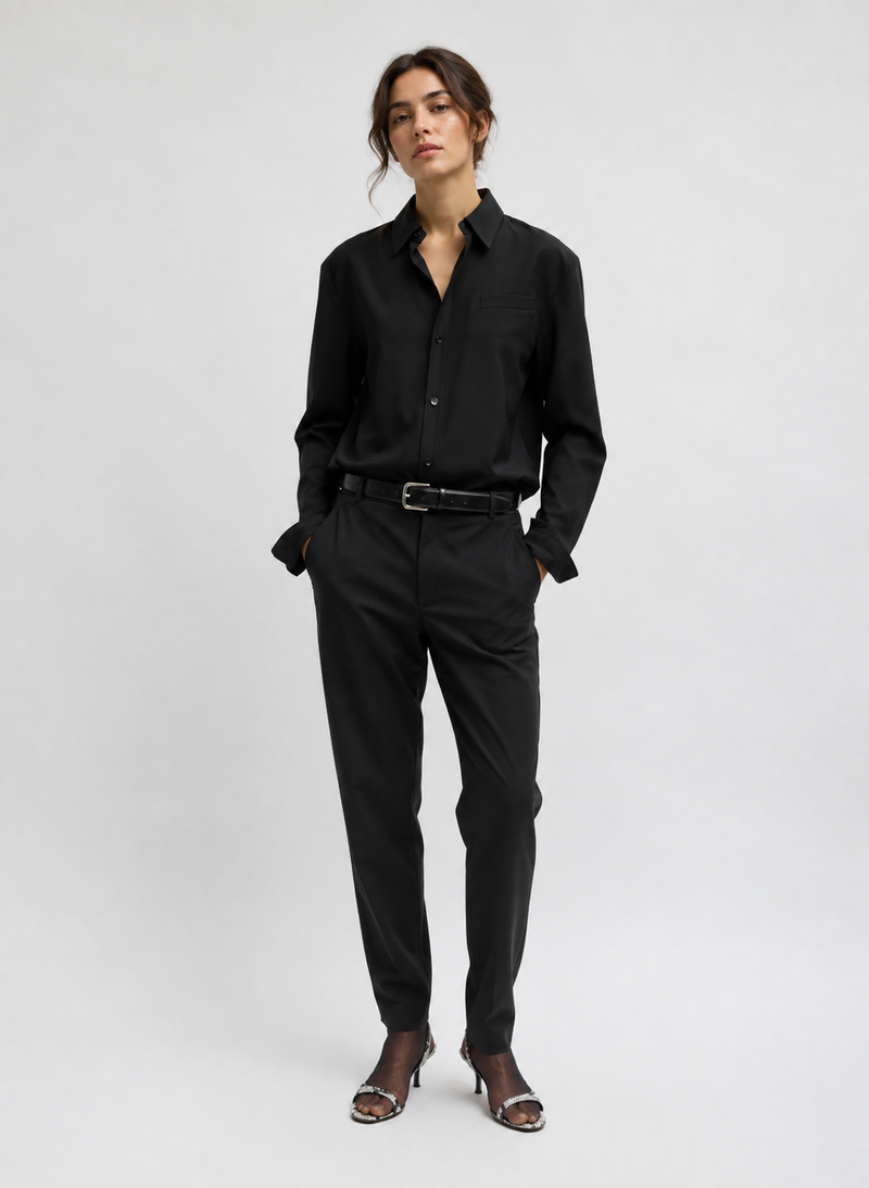 A model in a black long-sleeve button-up shirt and matching black trousers with a black belt, paired with brown heeled sandals by Tibi. 