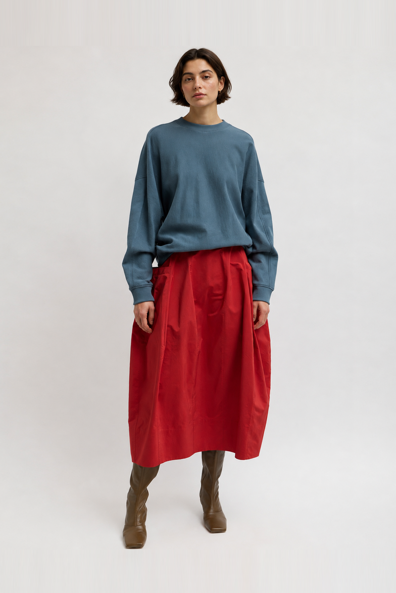 A model wearing a dusty blue long-sleeve crewneck sweatshirt, a bright red midi skirt, and brown ankle boots by Tibi. 