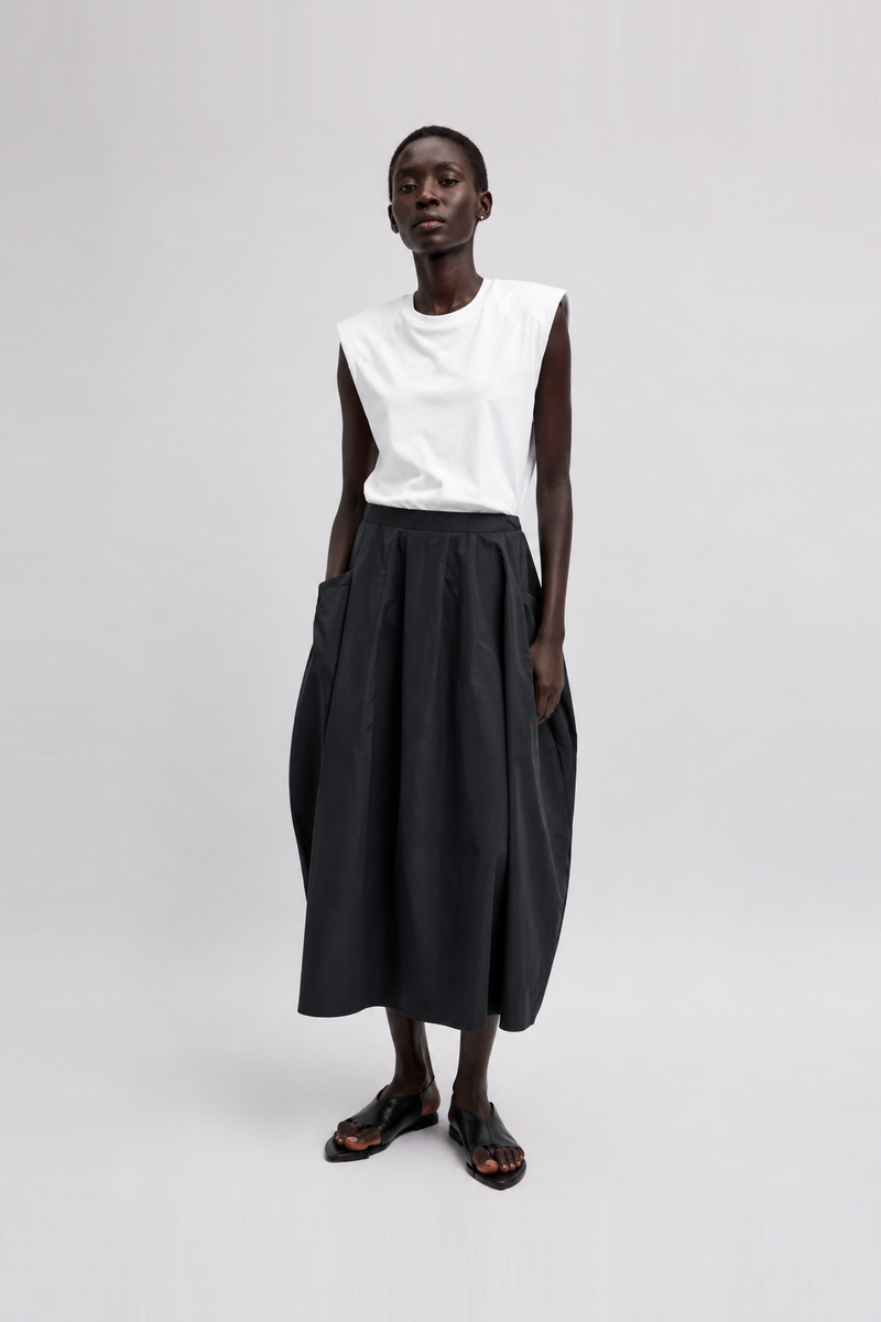 A model wearing a sleeveless white top and a voluminous black midi skirt with side pockets, paired with dark brown sandals from Tibi. 