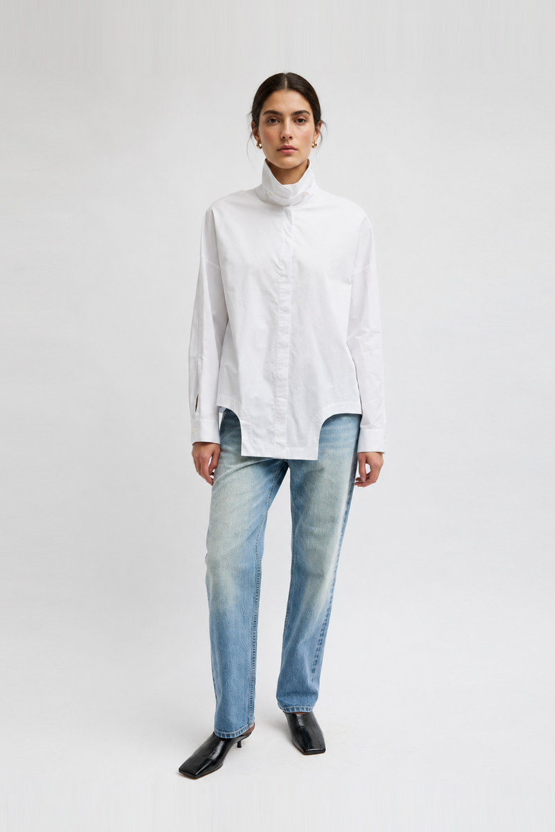 A model wearing a white long-sleeve mock-neck shirt, light-wash blue jeans, and black pointed shoes by Tibi. 