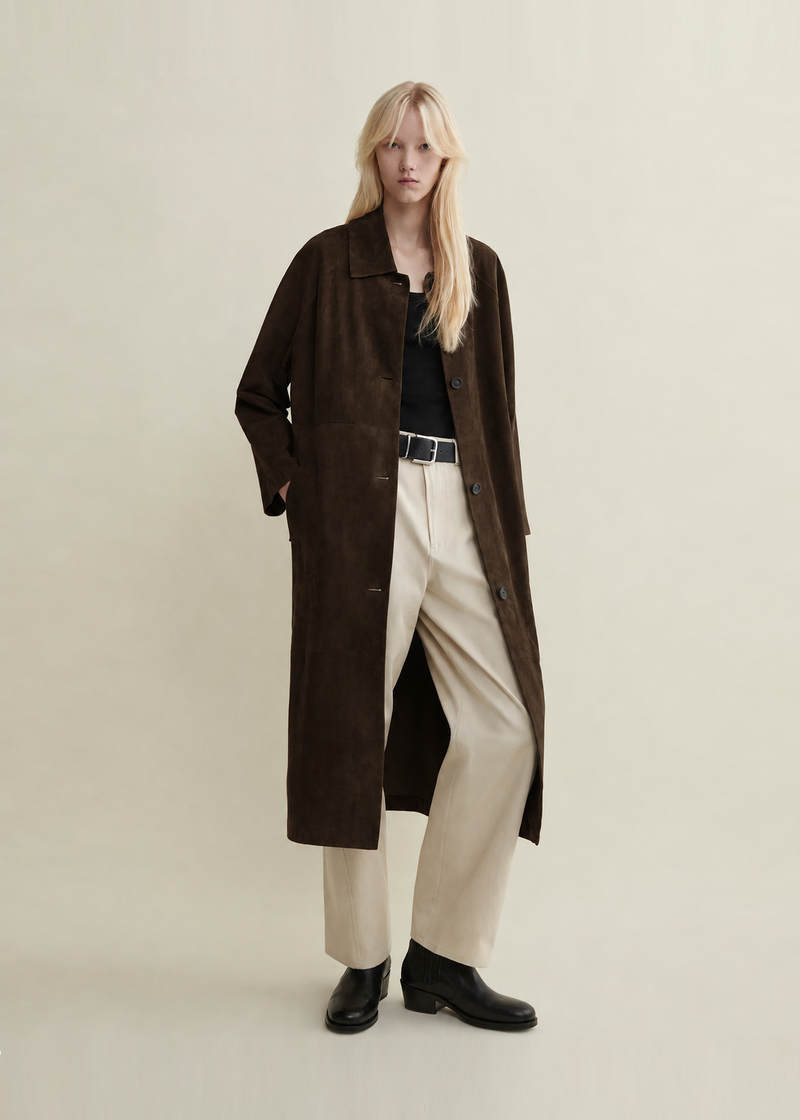 A model wearing a long dark brown suede-like coat over a black top and cream wide-leg trousers with black ankle boots from Arma. 