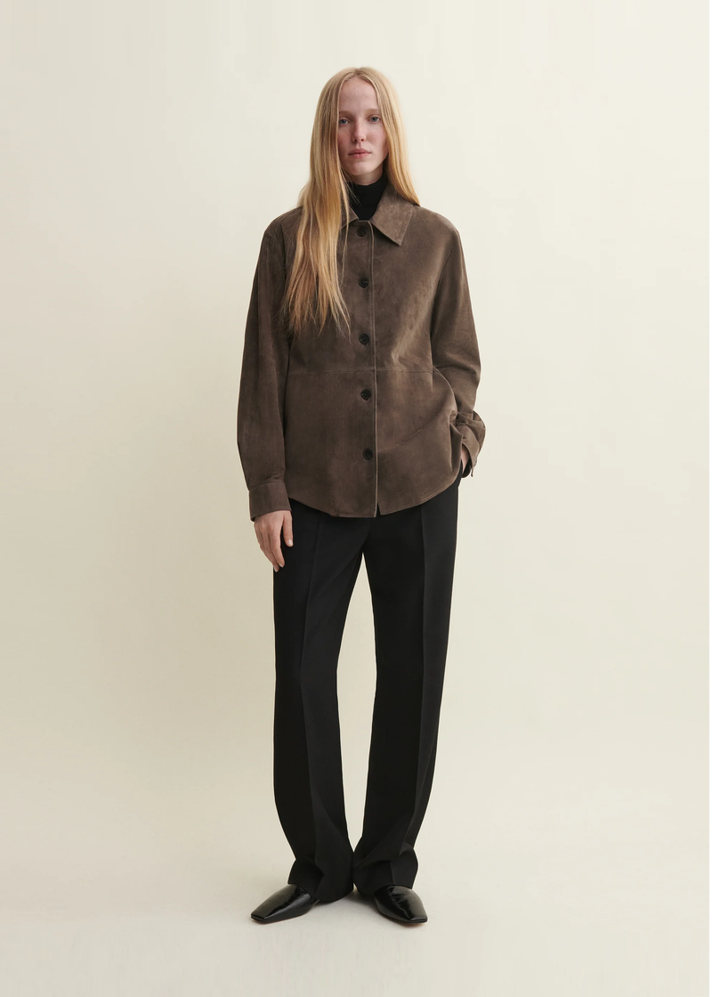 A model wearing a brown suede-like button-front jacket over a black turtleneck, black wide-leg trousers, and black shoes from Arma. 