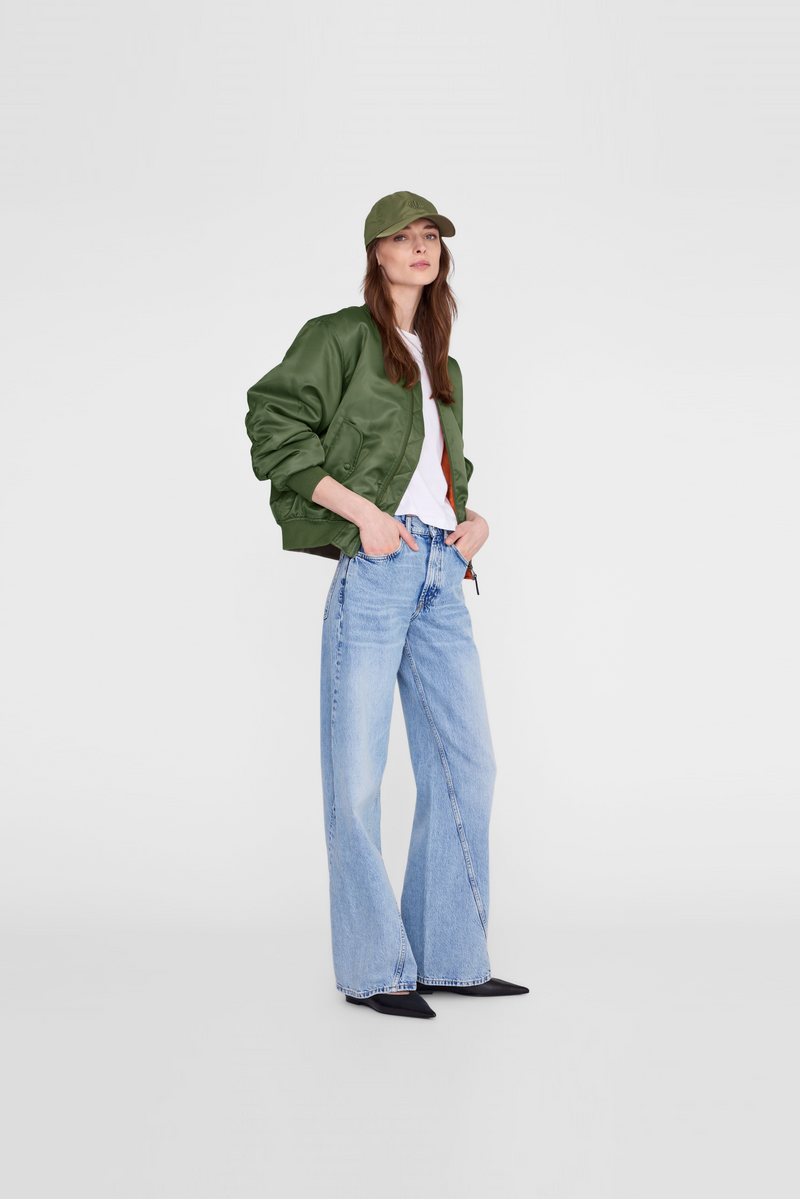 A model  wearing a green bomber jacket, light blue wide-leg jeans, a white top, black shoes, and a green cap by Anine Bing. 