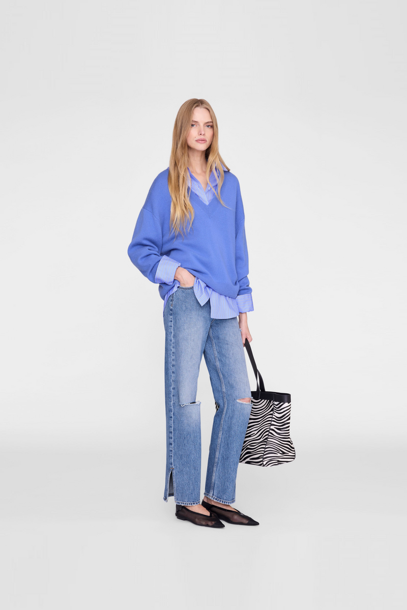A model wearing a bright blue knit sweater over a light blue collared shirt, light-wash blue jeans with knee patch details, and black flats by Anine Bing. 