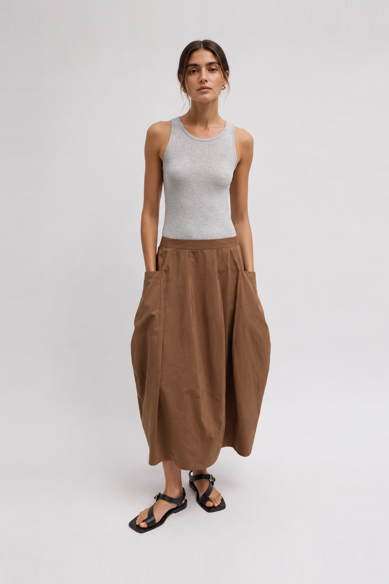 A model a light gray ribbed tank top and a high-waisted, voluminous brown linen maxi skirt with side pockets, paired with black strappy sandals by Tibi. 