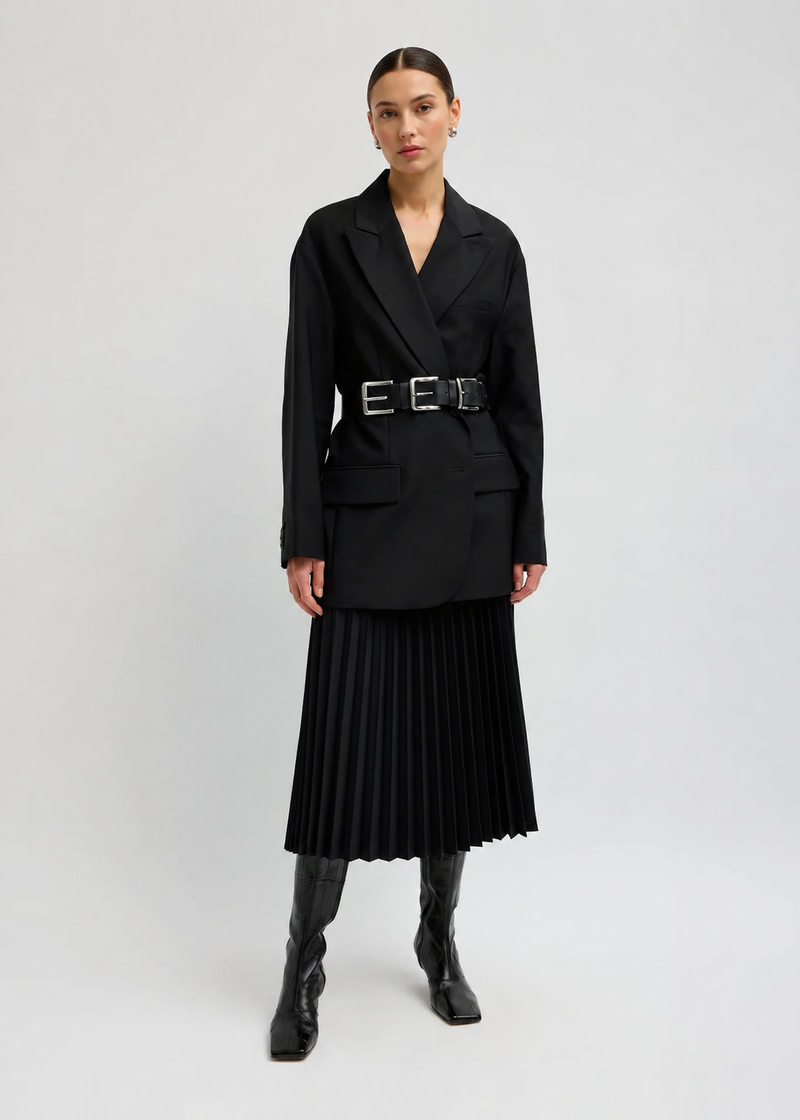 A model wearing a black long-sleeve blazer with a matching pleated midi skirt, a black belt with silver buckles, and black knee-high boots from Tibi.