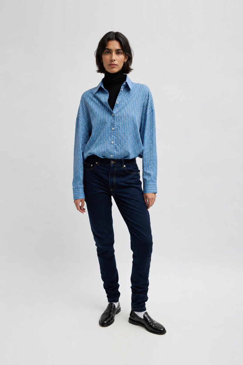 A model wearing a light blue and white striped button-down shirt, dark navy jeans, a black turtleneck, black loafers, and white socks by Tibi. 