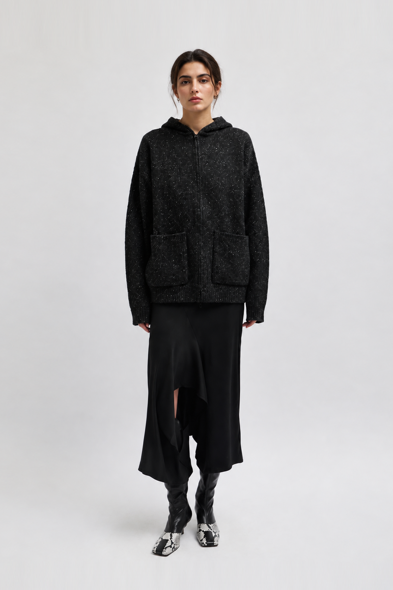 A model wearing a dark charcoal gray textured zip-up hoodie with front patch pockets, a long black skirt, and black boots with silver detailing from Tibi. 